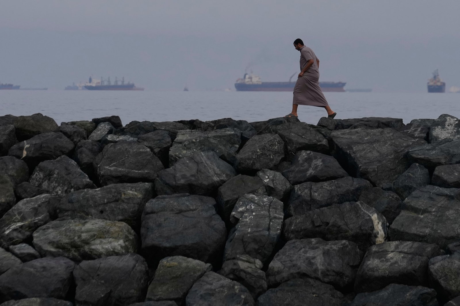 A man walks along the shore as oil tankers and cargo ships line up off the coast.