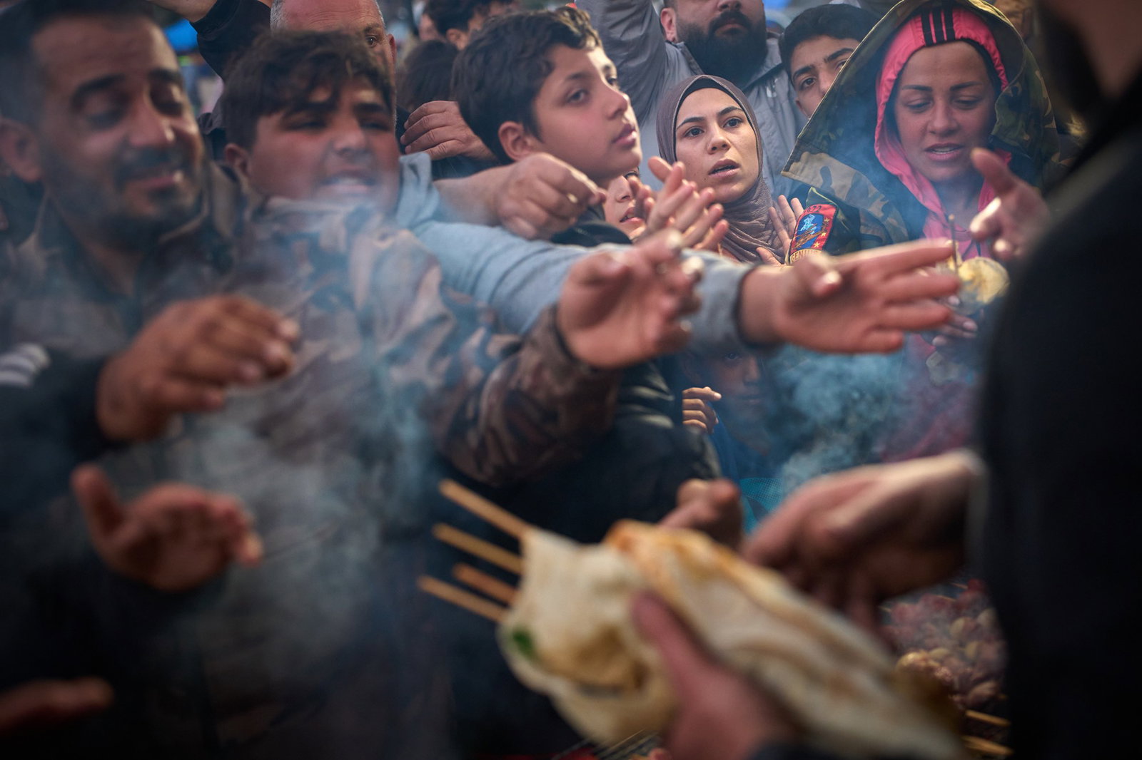 Displaced families extend their hands while waiting for donated food beside the tents they use as shelters