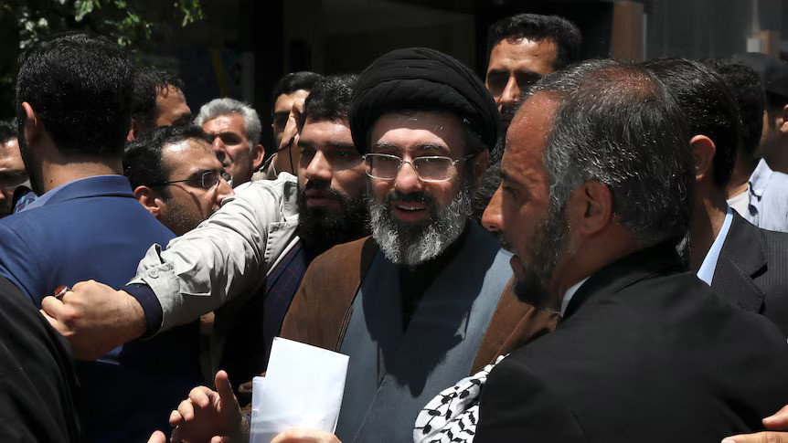 Mojtaba Khamenei, a middle-aged bearded man wearing glasses, stands among a crowd