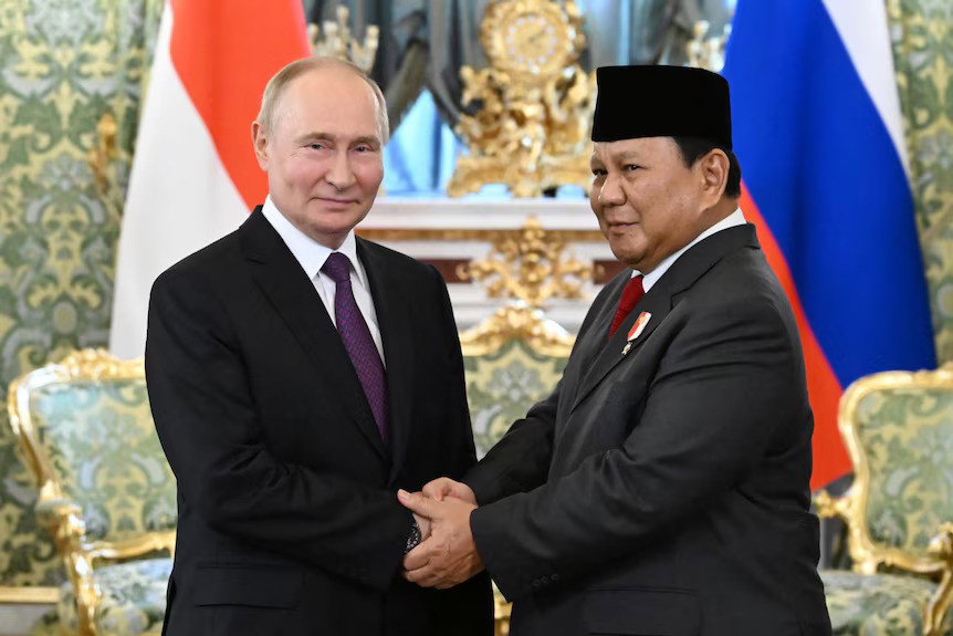 Vladimir Putin and Prabowo Subianto, both wearing suits, shake hands in front of the Russian and Indonesian flags