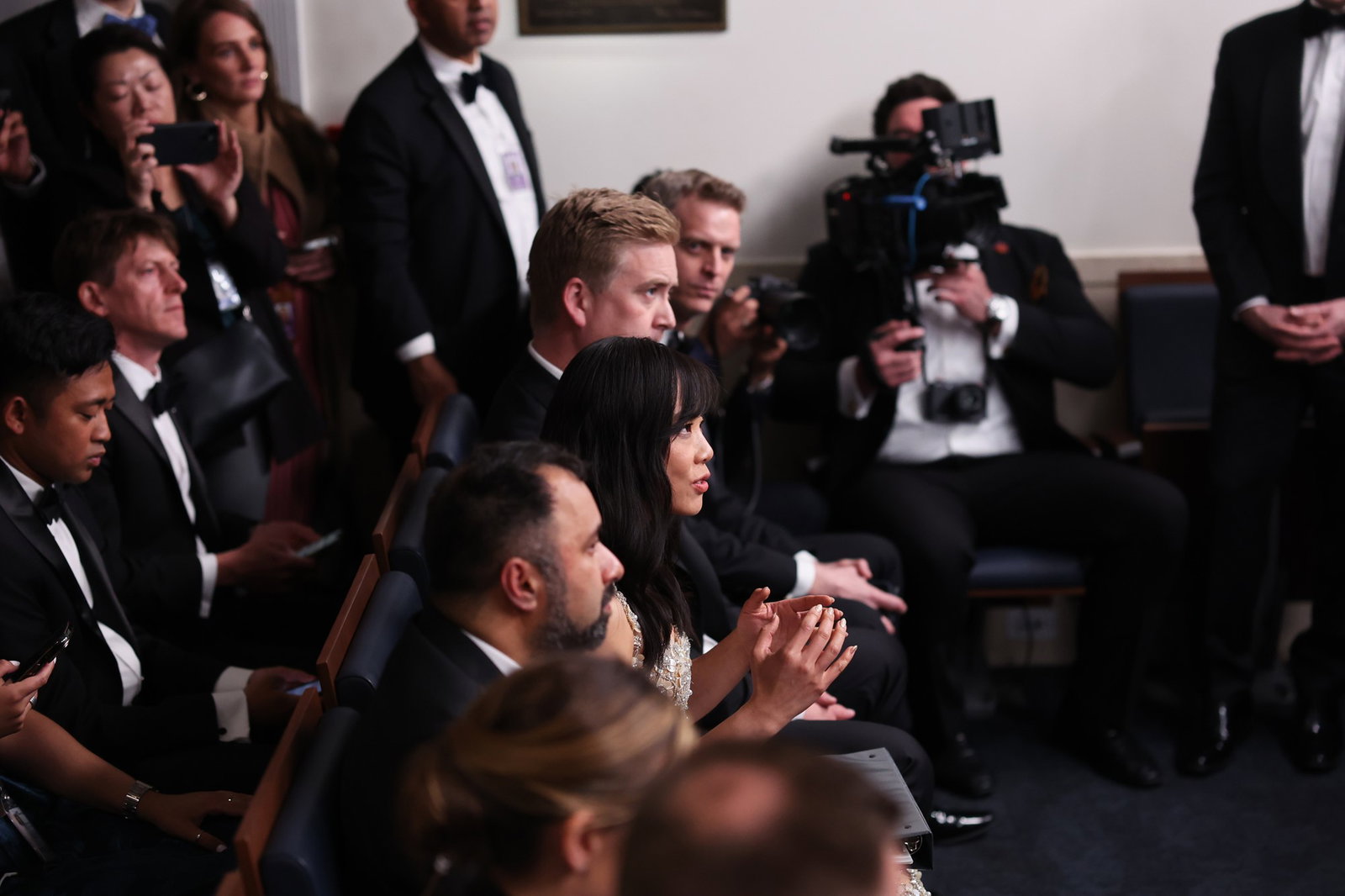 A middle-aged woman speaks while seated in a crowd of men in the White House press gallery.