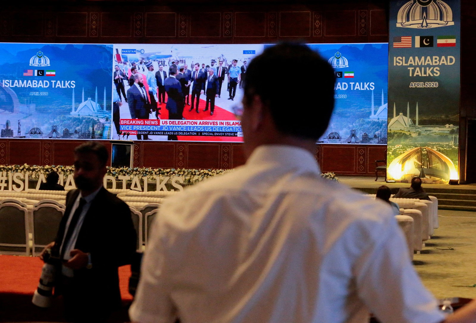 a man stands in the foreground, in the background is a big screen live streaming Vance's arrival in Pakistan