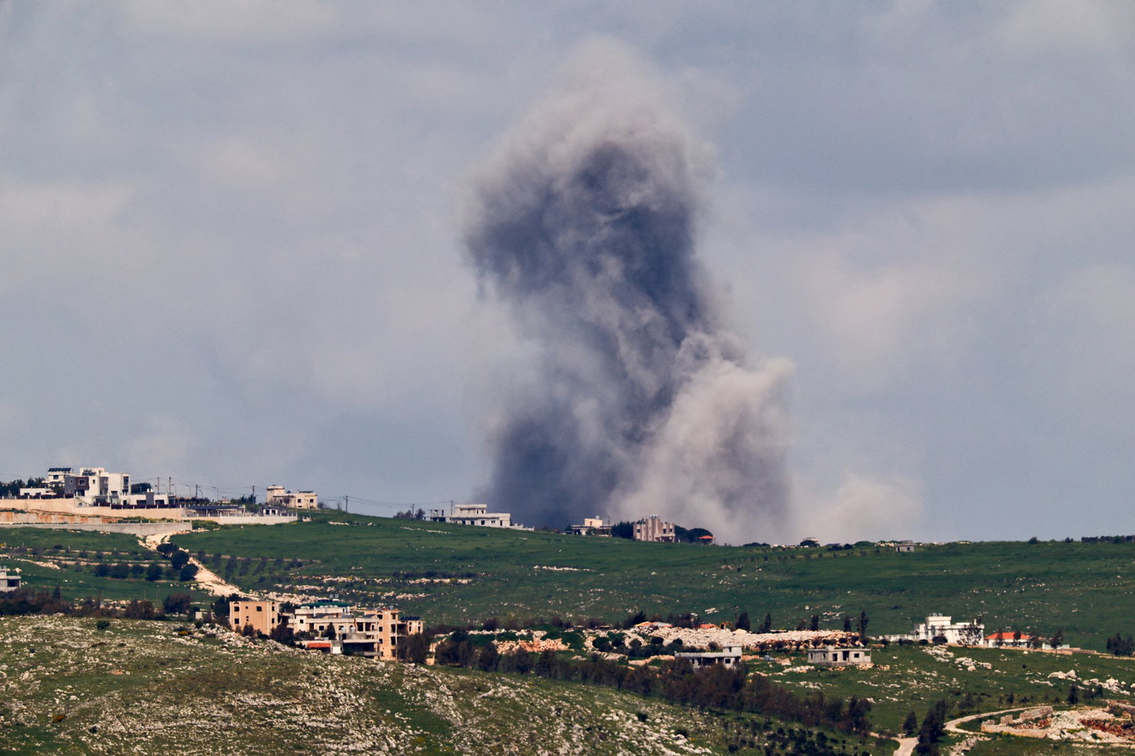 A ridge with buildings on it. A huge plume of smoke and dust rises from the otherside.