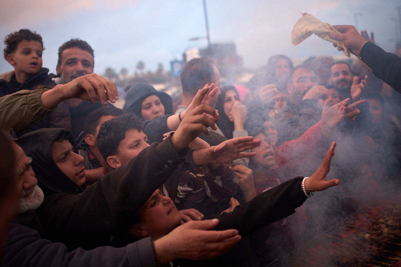 A crowd of men, women and children stretch their hands out as someone holds food out for them.