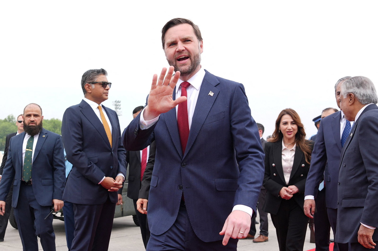 JD Vance wearing a dark blue suit with a white shirt and red tie. His hand is outsretch and his mouth is open. 