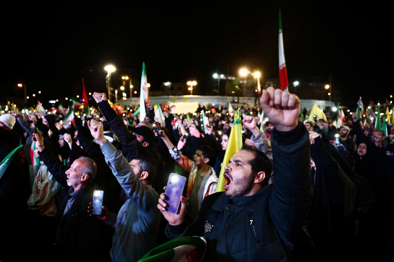 Men and women standing the dark holding Iranian white, green and red flags. Holding their fists in the air. 