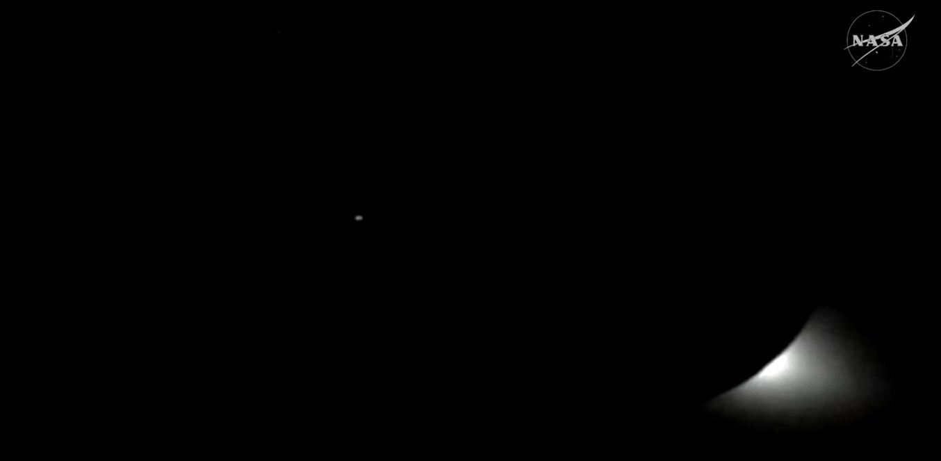 A small burt of white light peaks out from behind a black moon, in the black of space. To the left, a small dot (Earth) can be seen.