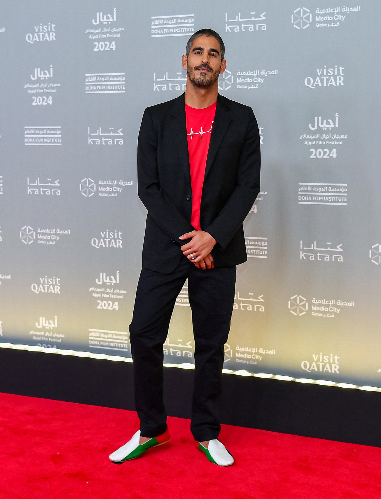 A man wearing a blazer, red shirt, dress pants and loafers on a red carpet in Qatar.