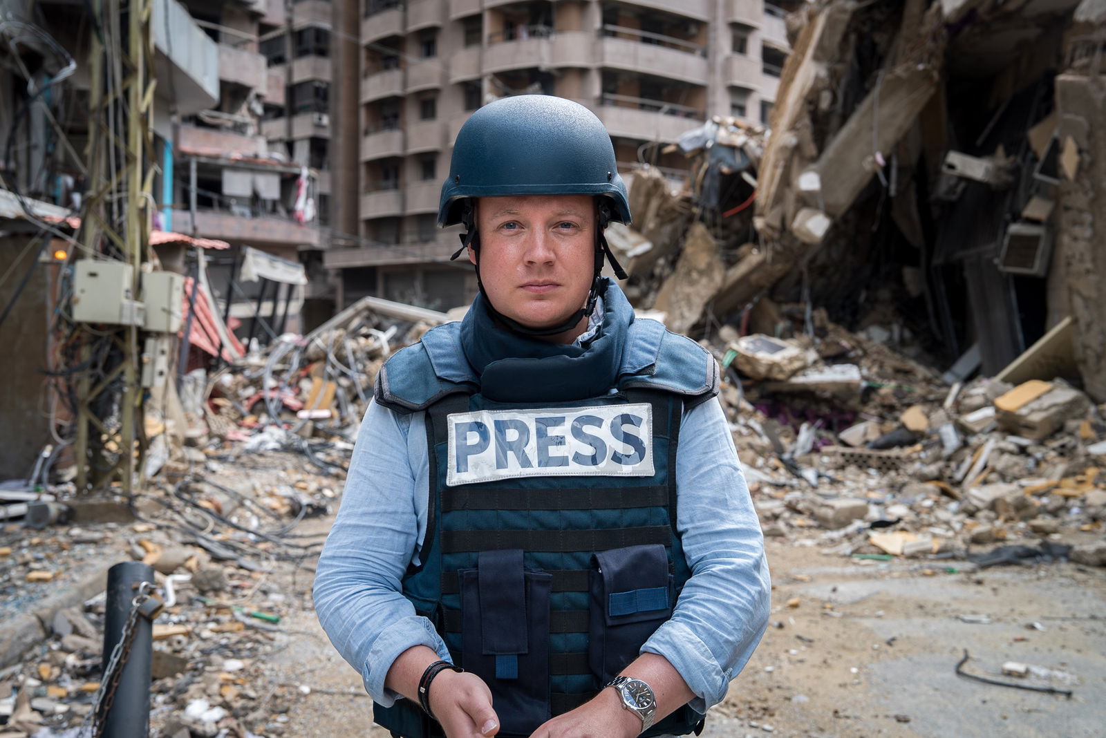 Matthew Doran stands in front of a destroyed street in a helmet and a bulletproof vest that says Press.