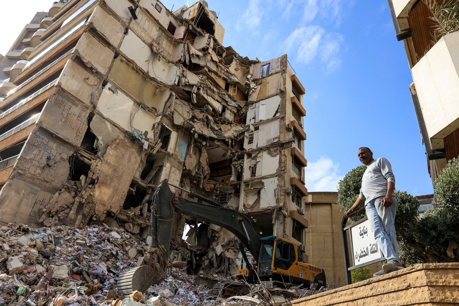 A man stands near a damaged building. A mechanical digger is situated in front of the building.