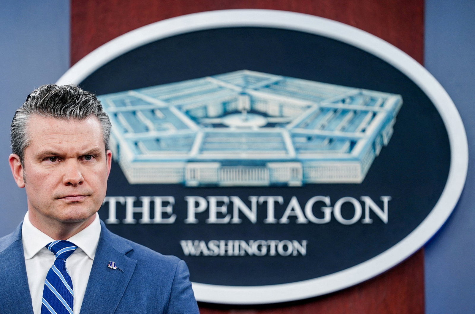 Pete Hegseth in a suit and tie standing in front of a logo for the Pentagon.