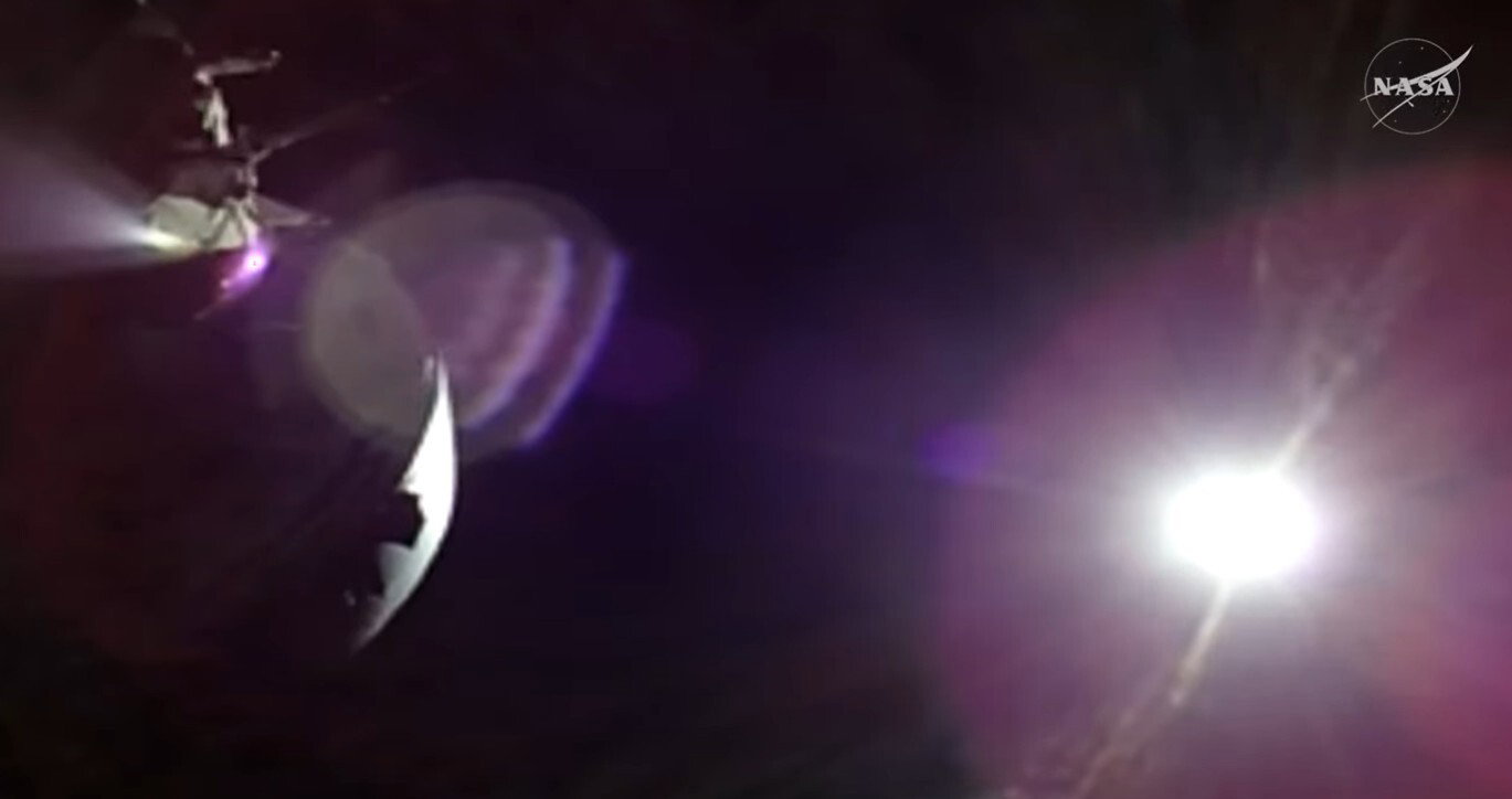 The sun is a bright white light in the darkness of space, casting a pink yellow sheen to the right of the image. To the left, part of a spacecraft in shadow can be seen.