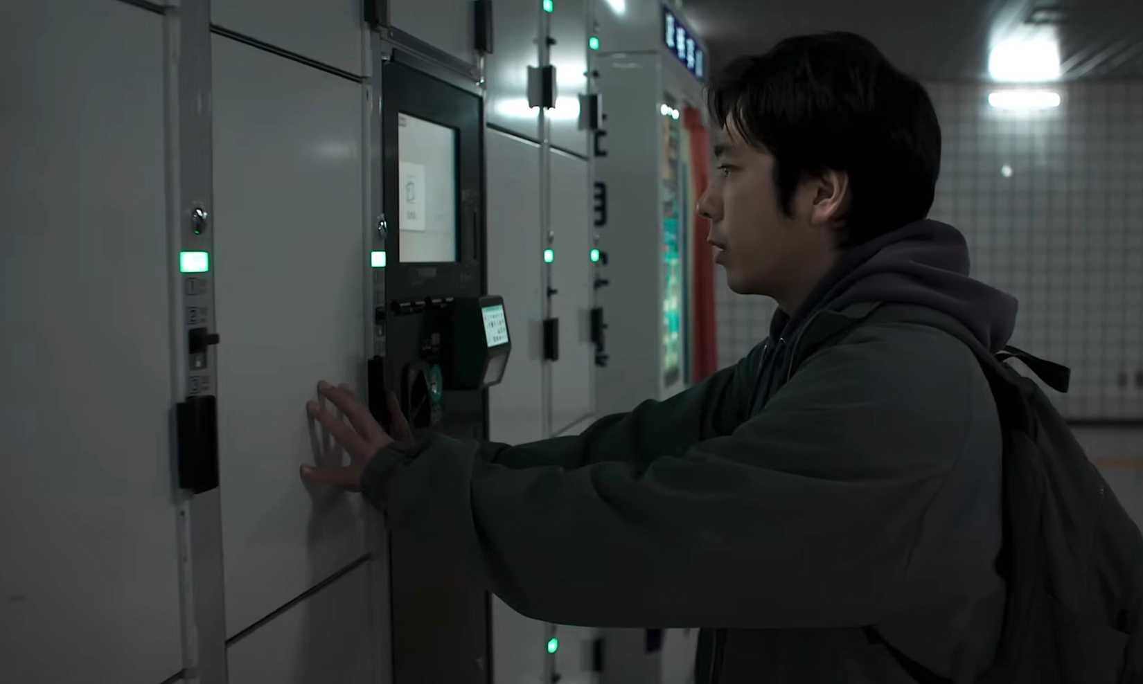 A man leans nervously against a bank of lockers next to a photo booth in a subway corridor in Exit 8