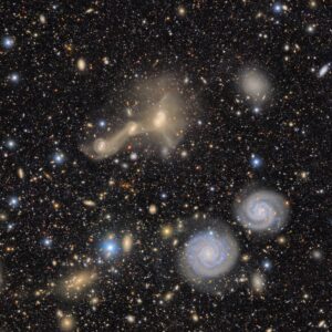 A telescope image is speckled with dozens-if not hundreds-of yellow, white and blue galaxies, some of which appear as small dots while others are larger and have a spiral shape similar to our own Milky Way. Just above the center of the image, several larger, yellow swirling galaxies appear to be pulling stars from one another.