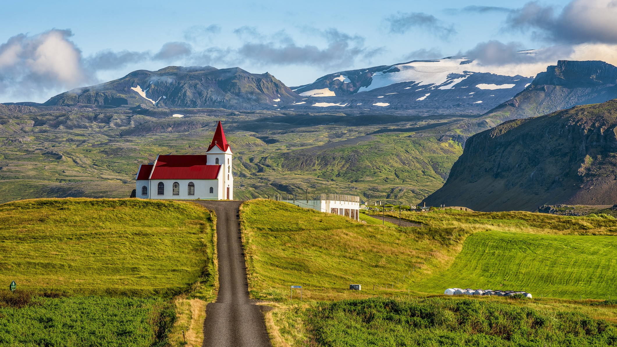 a small church with white walls and a red roof against a glacier backdrop. a road leads up to the scene.