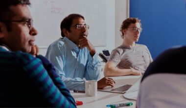 Krishna Gummadi, wearing rimless square glasses, a blue-and-white striped shirt, and with his hand on his chin, at a team meeting. To his right sits a colleague with curly hair, glasses, and a beige T-shirt. The photo was taken from the background, so two other colleagues can be seen from behind and in half-profile.