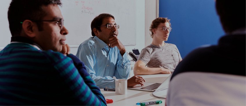 Krishna Gummadi, wearing rimless square glasses, a blue-and-white striped shirt, and with his hand on his chin, at a team meeting. To his right sits a colleague with curly hair, glasses, and a beige T-shirt. The photo was taken from the background, so two other colleagues can be seen from behind and in half-profile.