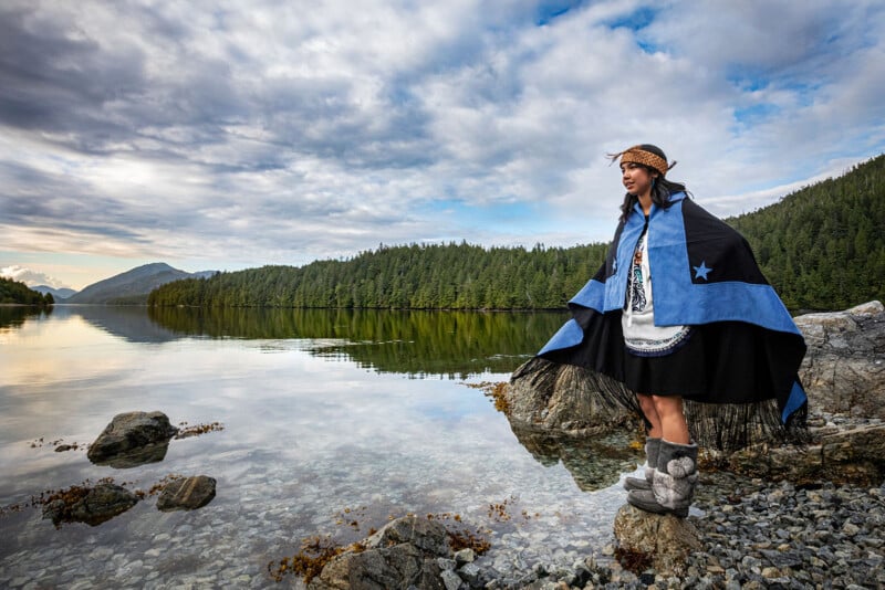 A person in traditional Indigenous clothing stands on a rock beside a calm lake, surrounded by forested hills and a cloudy sky, with mountains in the distance.