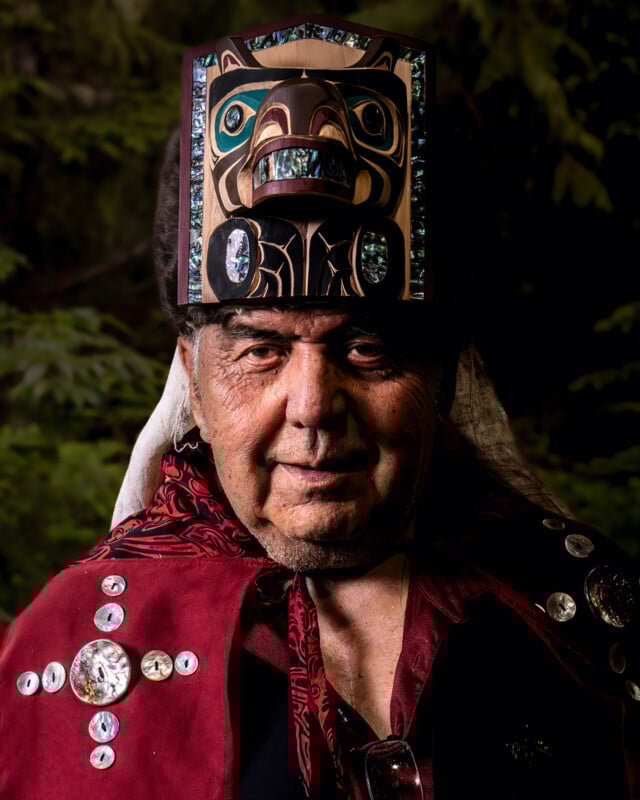 An elder wearing a traditional robe adorned with decorative buttons and a carved, painted wooden headdress stands in front of a blurred, leafy background.