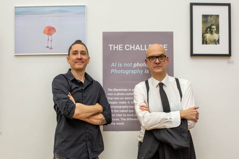 Two men stand with arms crossed in front of a wall with art and a sign about AI in photography. One man is casually dressed; the other wears a shirt, tie, and glasses. Framed photos hang on either side of the sign.