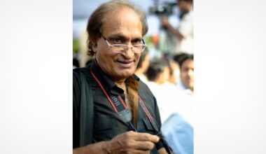 An older man with glasses holding a camera looks at the viewer and smiles. He is wearing a black shirt and a camera strap around his neck, with blurred people and outdoor activity in the background.