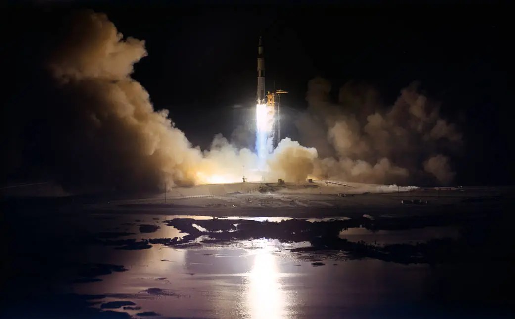 A rocket launches at night, emitting bright flames and smoke, its reflection visible in the water below—a powerful symbol of hope as Artemis II sets out on its journey from Earth to the moon.