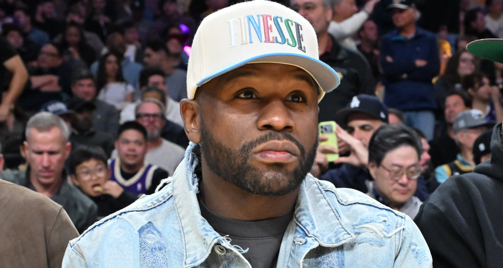Floyd Mayweather, in a denim jacket and cap, sits in a crowded arena, watching an event.