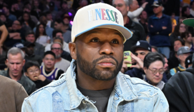 Floyd Mayweather, in a denim jacket and cap, sits in a crowded arena, watching an event.