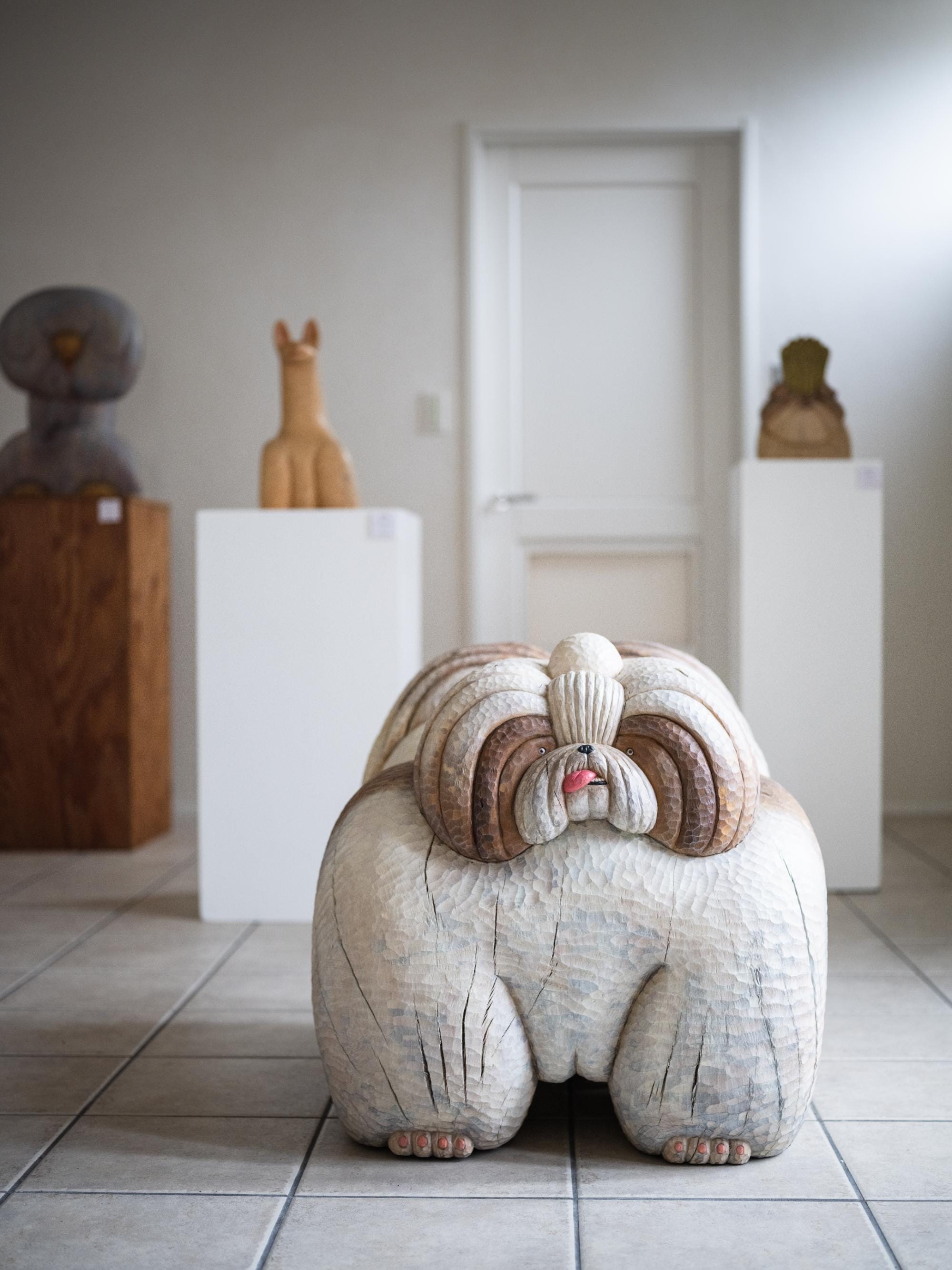 a wood-carved dog sculpture by Misato Sano stands on the ground. more wood-carved dogs stand on pedestals behind it