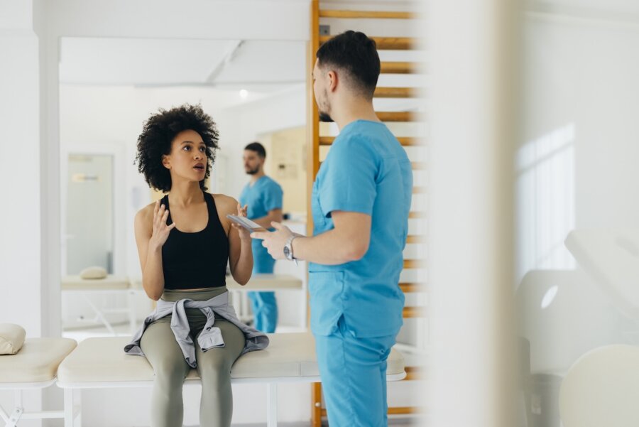 What the HECK is NAD? (And Should You Be Taking It?) - 2 A woman sits on an exam table in a medical clinic, talking to a healthcare professional in blue scrubs who is holding a clipboard and discussing the potential benefits of NAD supplements.