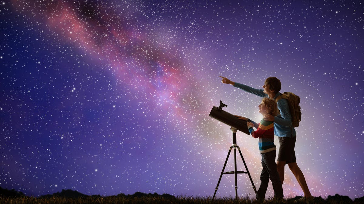 Two people with a telescope look up into a night sky that's full of stars and the Milky Way.