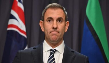 Australian federal treasurer Jim Chalmers at a press conference in Canberra. The image is a medium shot. Chalmers wears a black suit with striped blue tie and a wan expression on his face. Behind him are the Australian and Torres Strait Island flags.