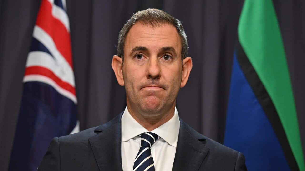 Australian federal treasurer Jim Chalmers at a press conference in Canberra. The image is a medium shot. Chalmers wears a black suit with striped blue tie and a wan expression on his face. Behind him are the Australian and Torres Strait Island flags.
