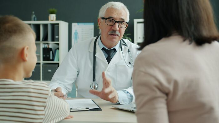 Doctor with glasses and white coat explaining concerning hospital truths to a woman and child in a medical office.
