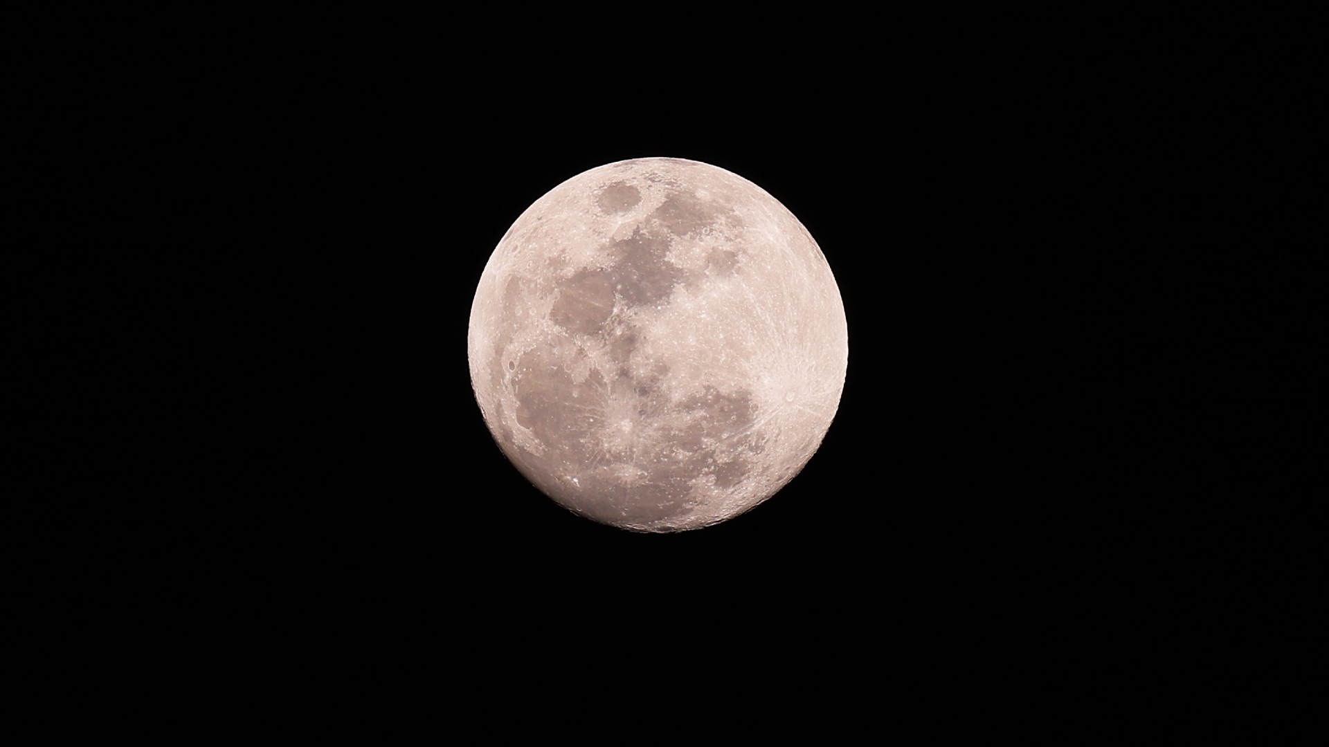 A silvery full moon is pictured glowing in a black night sky. Dark lunar seas and bright young craters can be seen marking its surface.