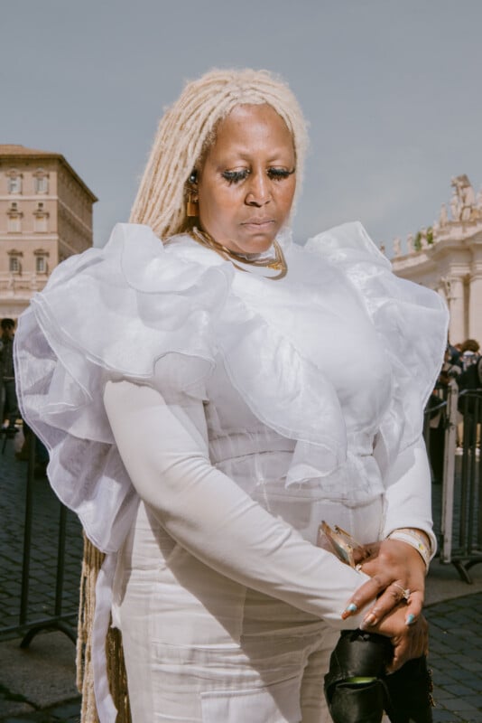 A woman with long blonde dreadlocks wears a dramatic white ruffled dress, standing outdoors in a city square on a sunny day, with historic buildings and a blue sky in the background.