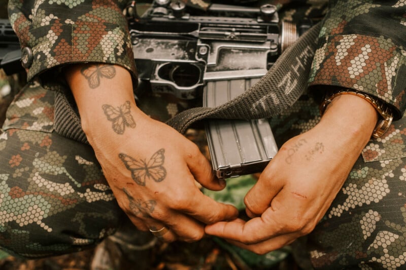 A person in camouflage uniform with butterfly tattoos on their hands holds a rifle across their lap, showing details of the tattoos, gold bracelet, and rifle magazine.