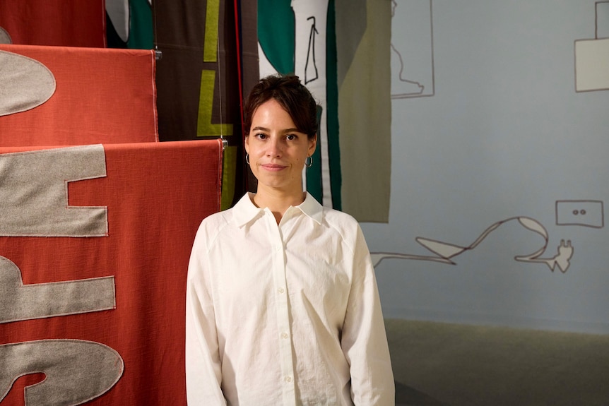 A young woman with short dark hair, wearing a white button-up shirt, standing in front of colourful fabric banners.