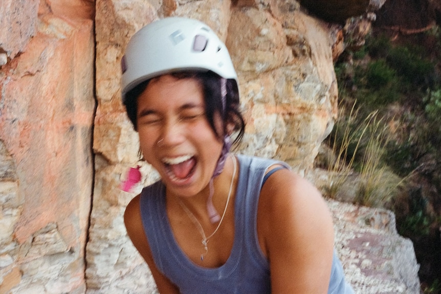 Woman laughing with mouth wide open, eyes shut, with white climbing helmet on in front of rock outdoors