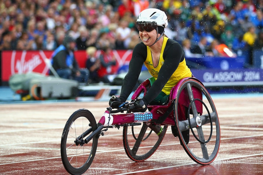 Angie Ballard, in helmet and yellow and green racing uniform, sits in racing wheelchair with wide smile, poking out tongue.