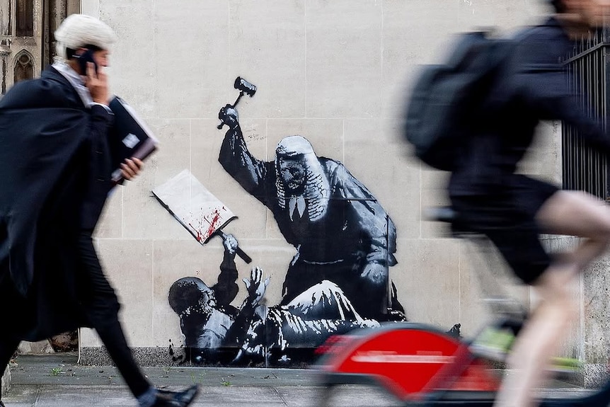 People walk in front of an artwork on a wall of a judge beating a protester with a gavel