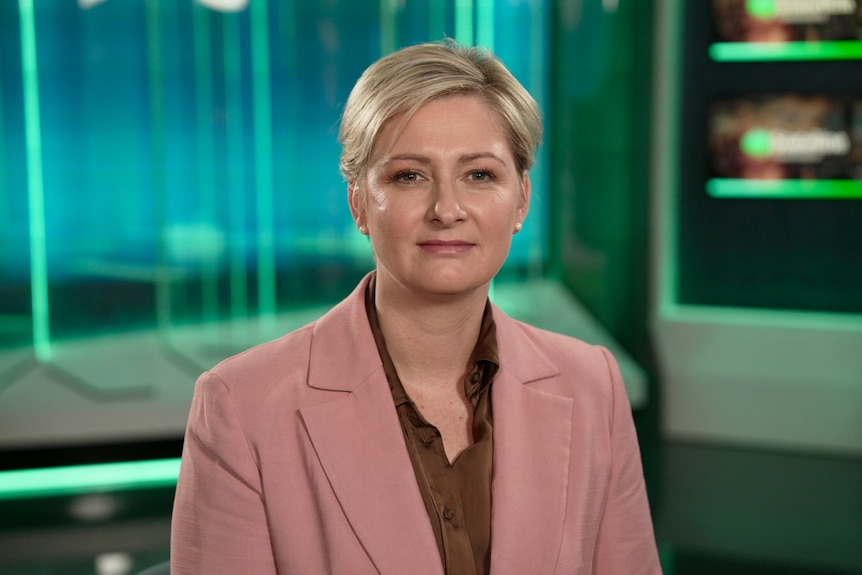 A woman in a pink jacket staring directly at the camera. A blue and green lit studio in the background.