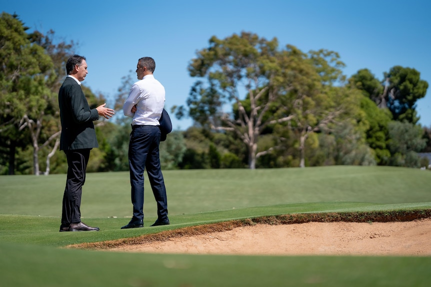Two men in suits are standing on a golf course near a sand bunker.