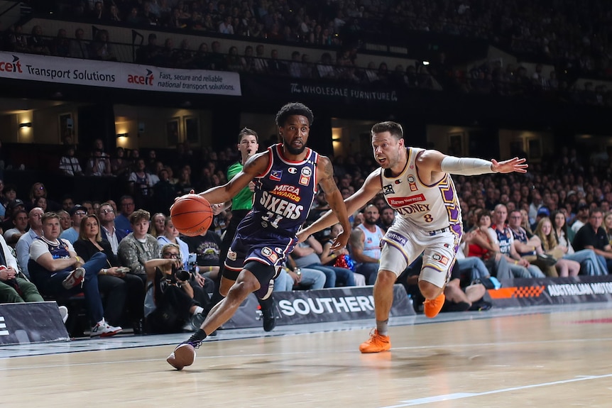 Two basketball players in action, one in a white uniform and one in a navy uniform.