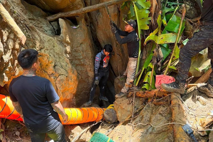 Three men in helmets stand at the muddy entrance to a small cave.