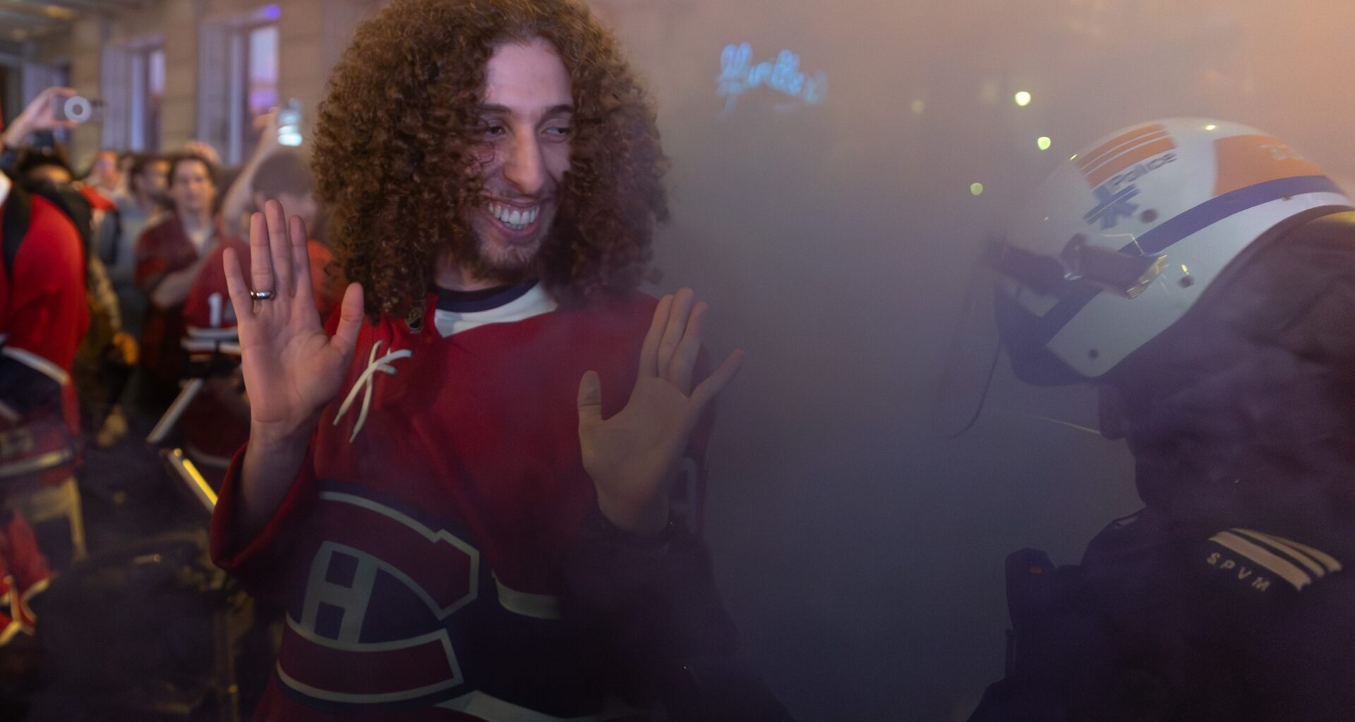 A man in a Canadiens jersey smiles at a police officer in riot gear as they're surrounded by smoke