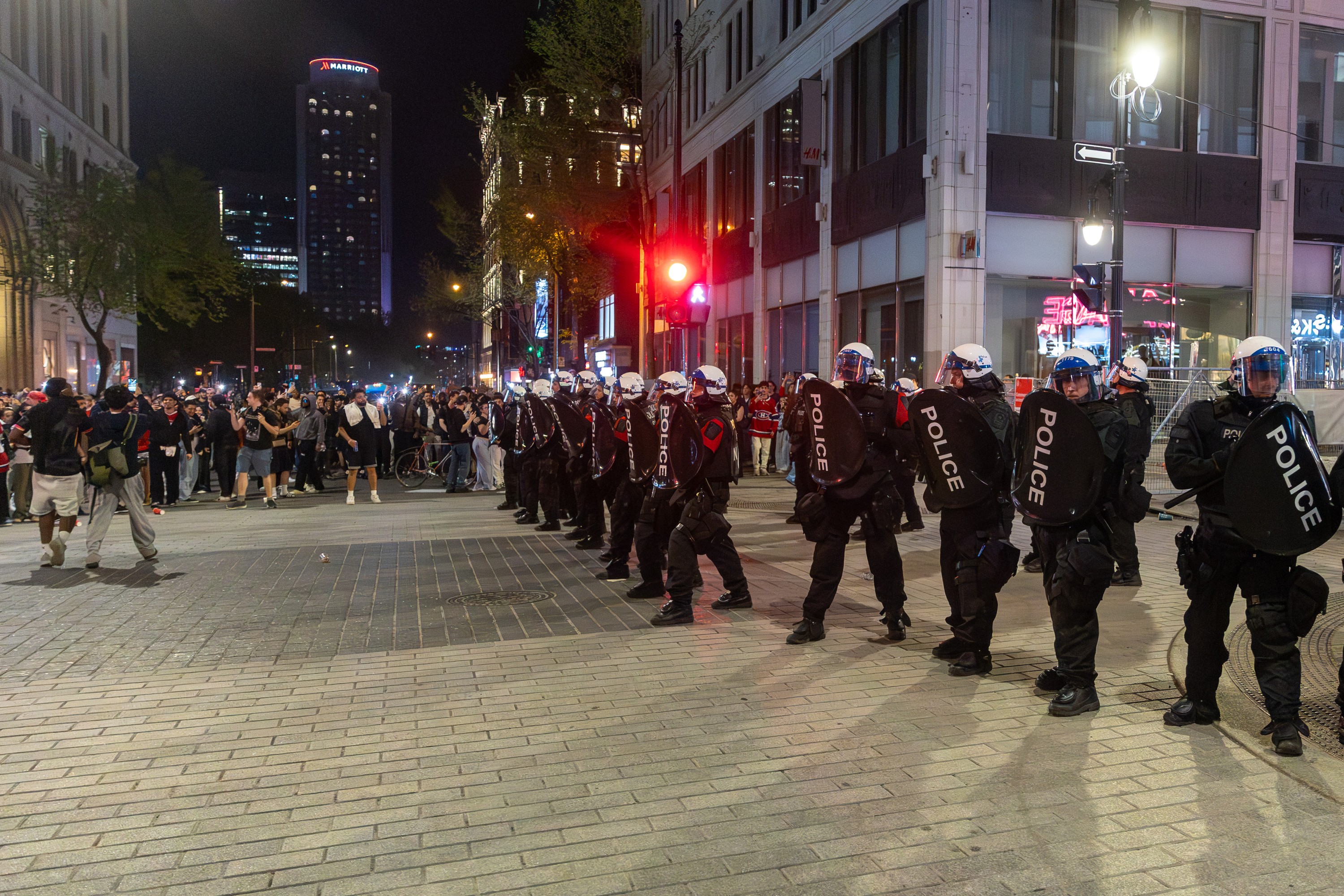 Police in riot gear line up at an intersection, with many civilians nearby.