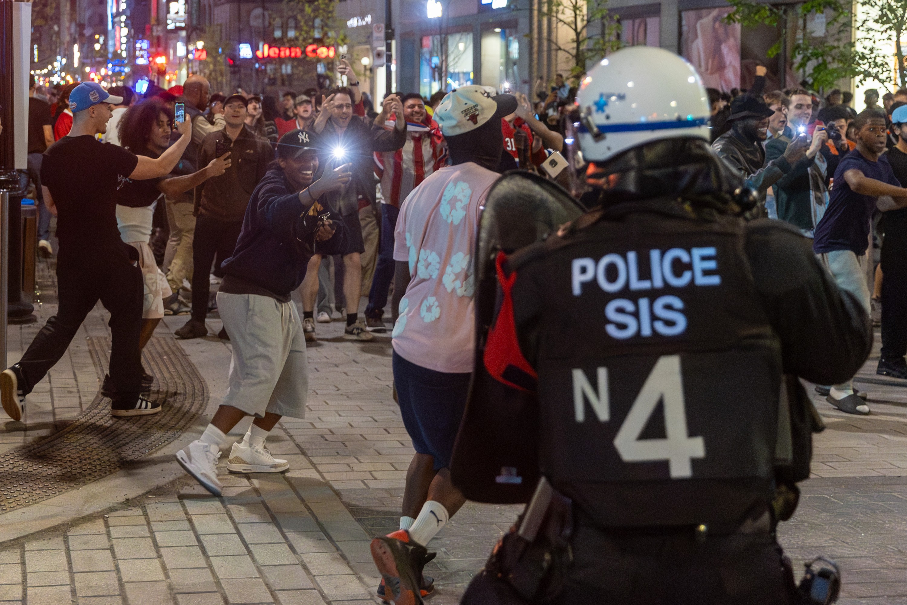 People gleefully record video on smartphones with a police officer in riot gear in the foreground on a downtown street at night.