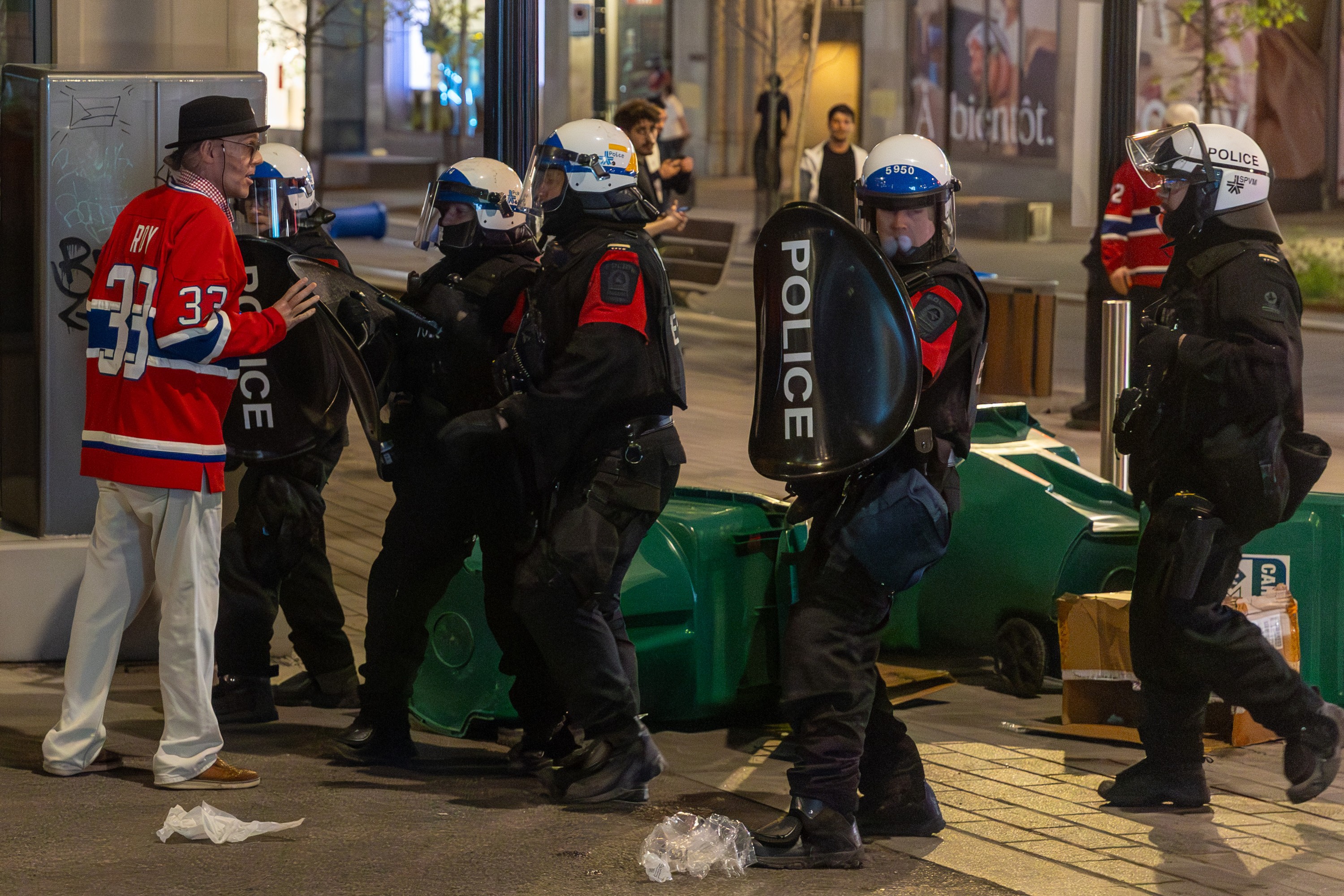 Police in riot gear interact with a man in a Canadiens jersey on a downtown street, with overturned recycling bins behind the police.