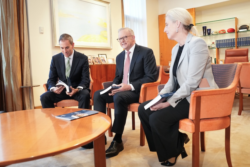 Jim, Anthony and Katie sit around a round table, Jim looks down, katie at the prime minister who is talking. Books behind.